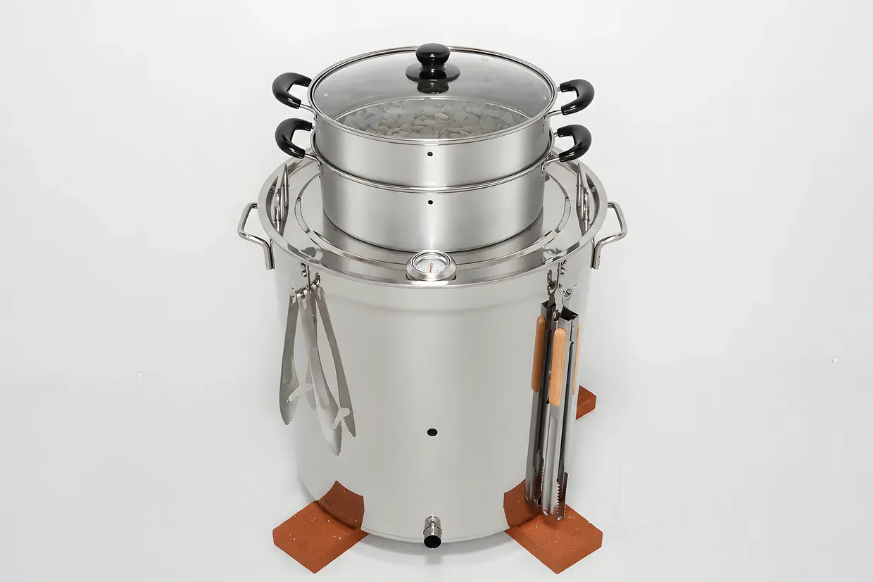 ステンレス製の焼き芋機の全体。上に蒸し器、脇に専用のトングと火ばさみ、足元にレンガ